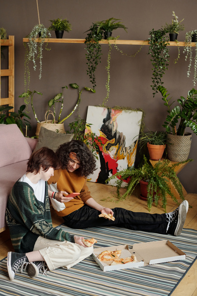 A girl with short hair in an oversize sweater light trousers and sneakers holding a piece of pizza in her hand is sitting on a striped carpet next to a guy in a bandana and a mustard-colored sweatshirt who shows her a photo on his phone